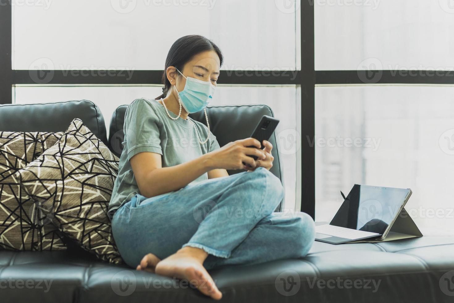 donna che indossa la maschera protettiva sedersi sul divano utilizzando lo smartphone in quarantena foto