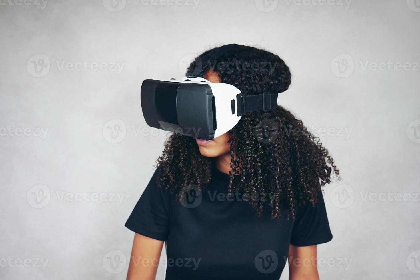 una bella giovane donna nera con i capelli afro ricci indossa l'auricolare vr di realtà virtuale e gioca ai videogiochi in studio con sfondo grigio foto