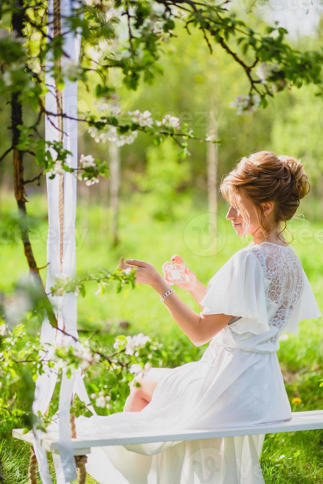 giovane sposa con capelli biondi in vestaglia bianca utilizzando profumo su un'altalena di corda foto