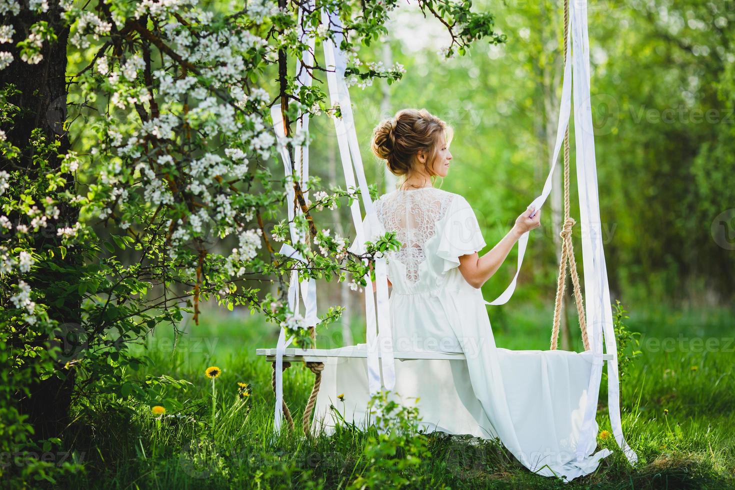 giovane sposa con capelli biondi in vestaglia bianca in posa su un'altalena di corda foto