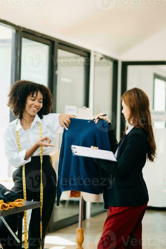 coppia di moda progettisti Lavorando con tessuto e capi di abbigliamento schizzi a il studio pieno di sartoria utensili e detiene tavoletta e il computer portatile. foto