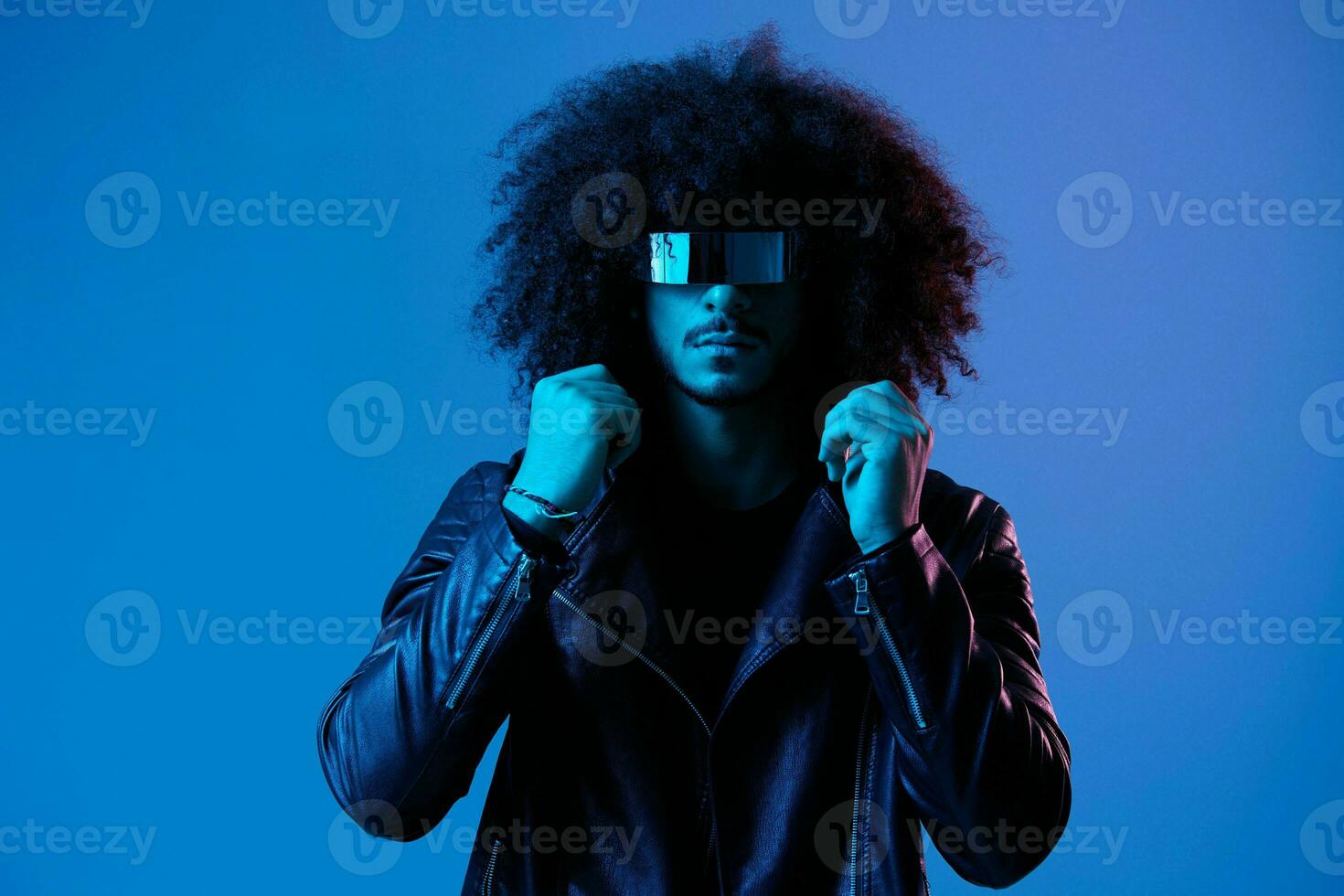 ritratto di moda uomo con Riccio capelli con elegante bicchieri su blu sfondo multinazionale, colorato luce, nero pelle giacca tendenza, moderno concetto. foto