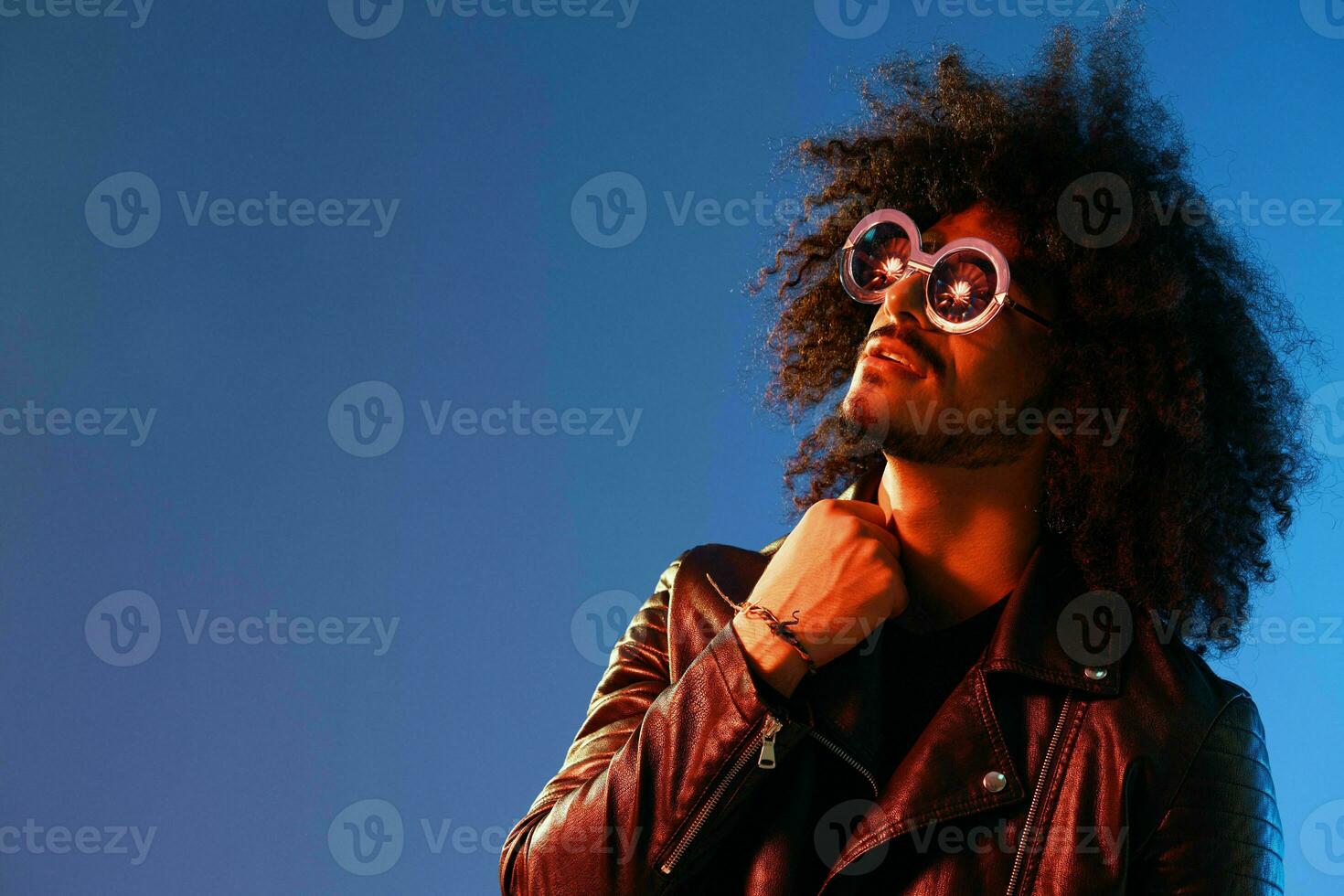 ritratto di un' elegante uomo con Riccio capelli con bicchieri Sorridi su blu sfondo multinazionale, colore luce, nero pelle giacca tendenza, moderno concetto. foto