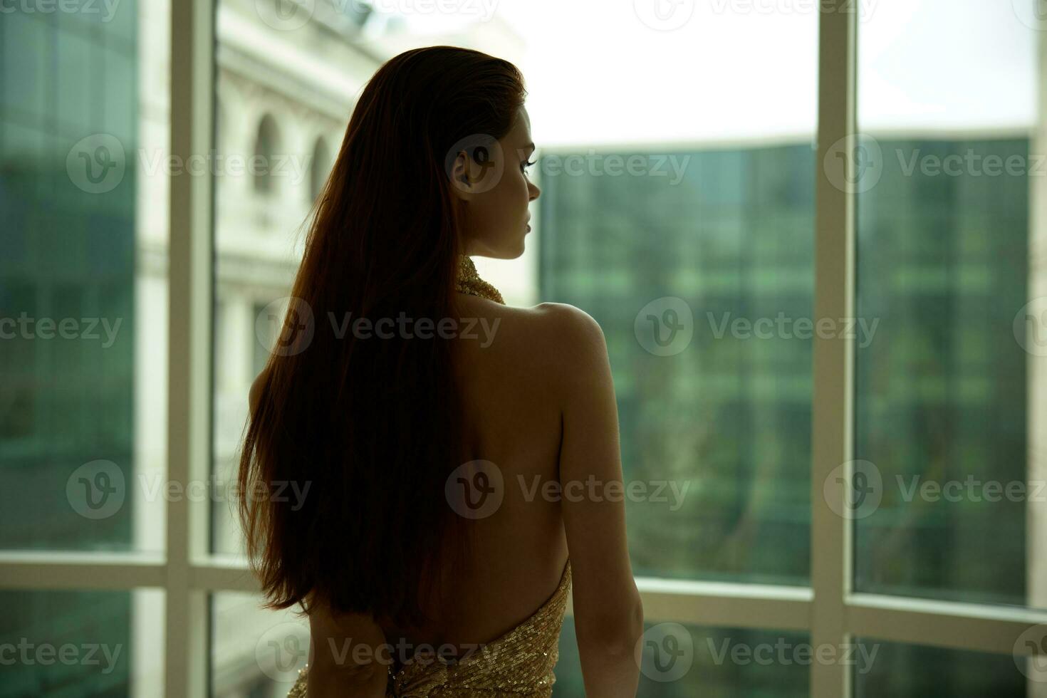 donna nel oro paillettes vestito in piedi di il finestra in posa e guardare a il città nel un' elegante Guarda con lungo capelli, progettista vestito con Aperto Indietro, festa Guarda foto