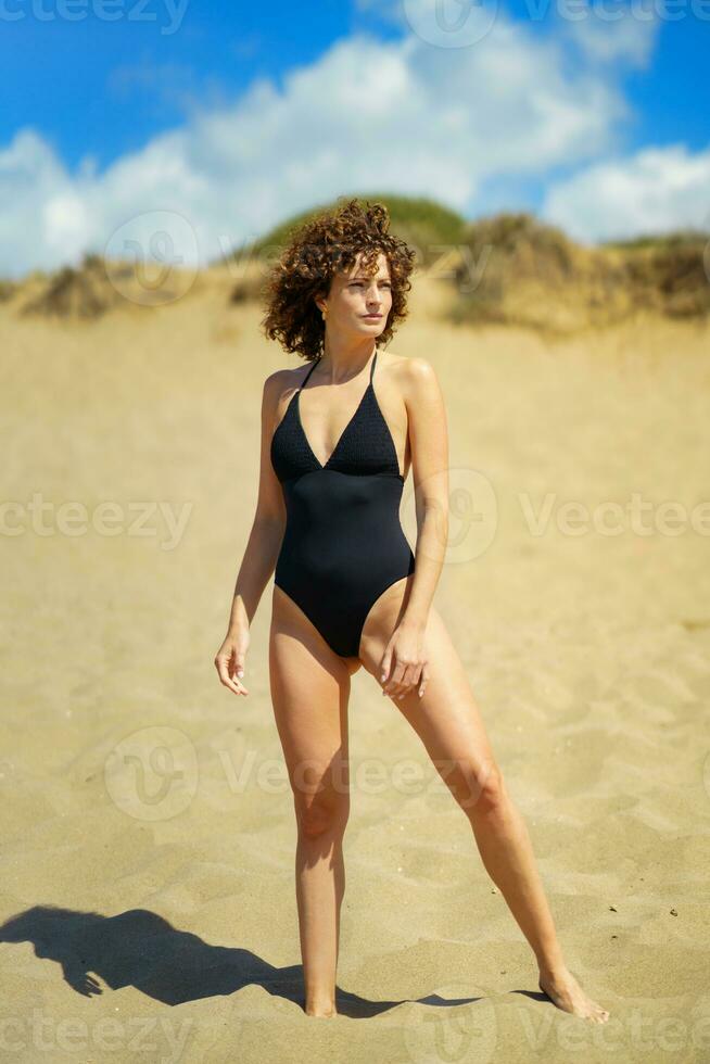 fiducioso donna nel costume da bagno in piedi su sabbioso spiaggia foto