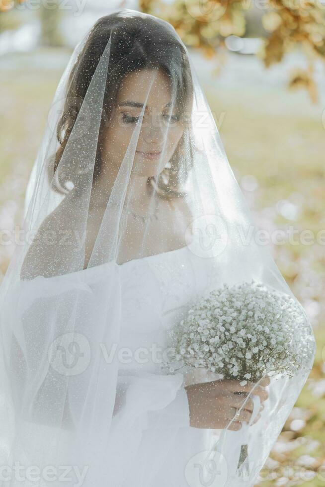 elegante bellissimo sposa sotto un' velo sta nel davanti e sembra a un' mazzo nel un' bianca nozze vestire. il sposa nel un' bellissimo luce. foto