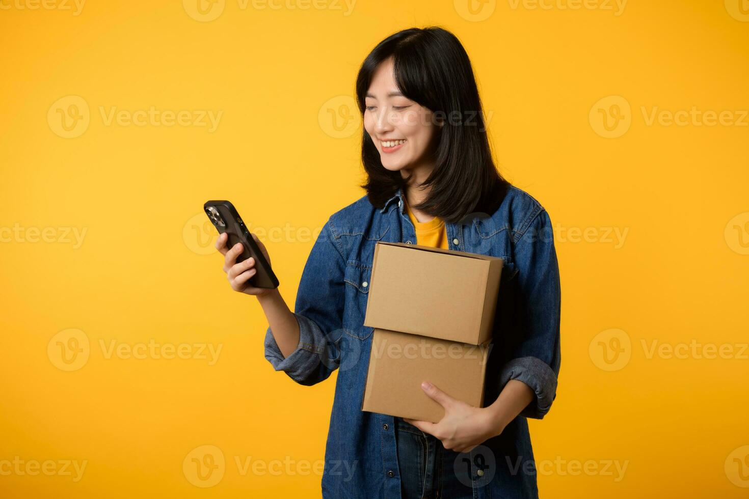 ritratto asiatico giovane donna indossare giallo maglietta e denim camicia Tenere mobile Telefono e pacco scatola isolato su giallo studio sfondo, consegna Corriere e spedizione servizio concetto. foto