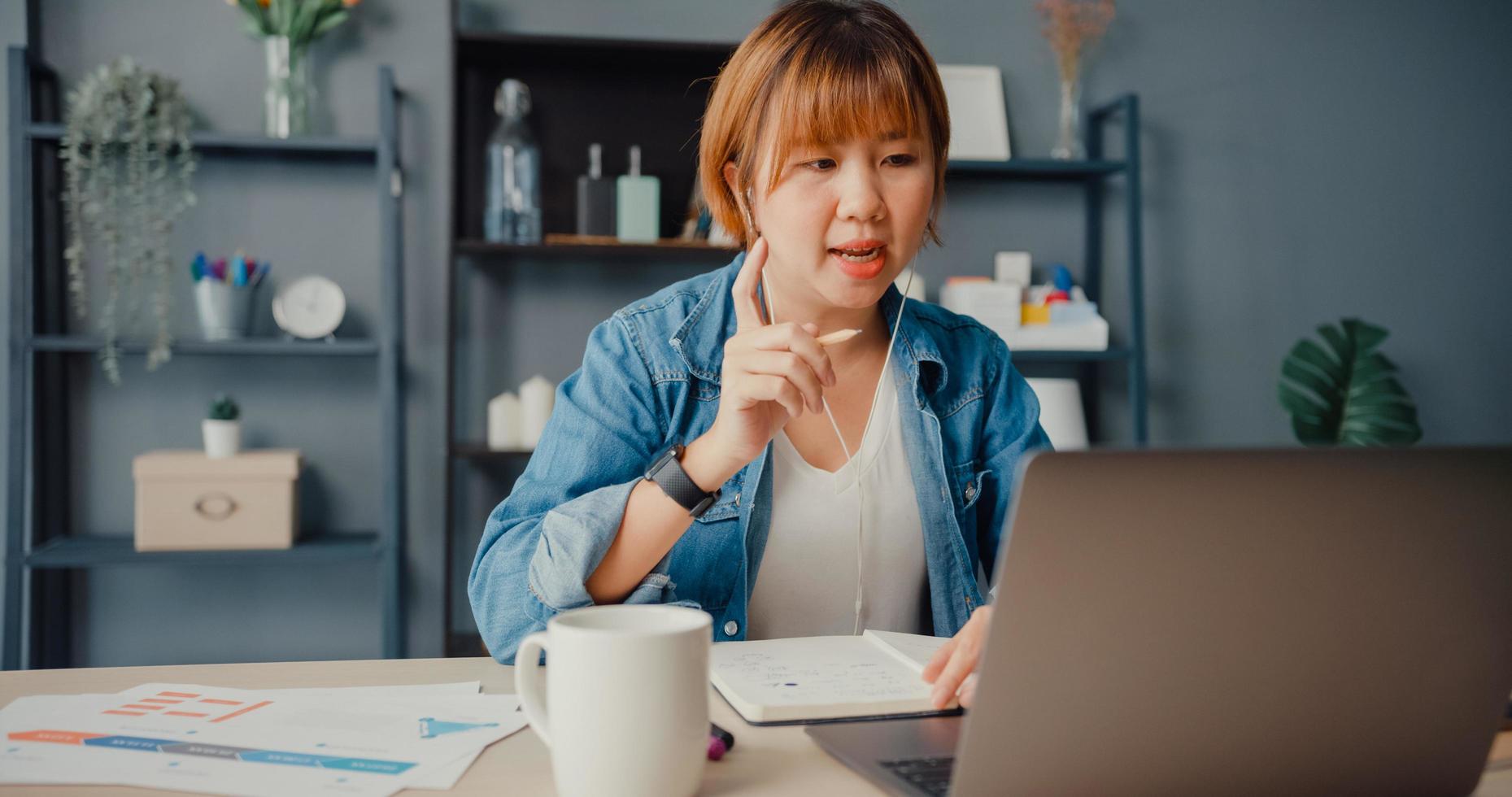 donna d'affari asiatica che utilizza laptop parla con i colleghi del piano in videochiamata mentre lavora da casa in soggiorno foto