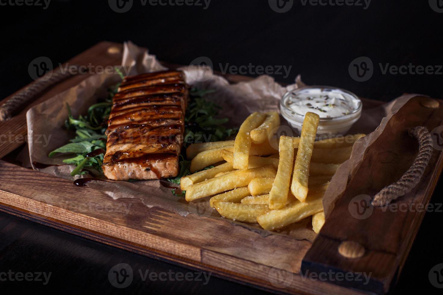 filetto di salmone con patatine fritte su un vassoio di legno, bella porzione, sfondo scuro foto