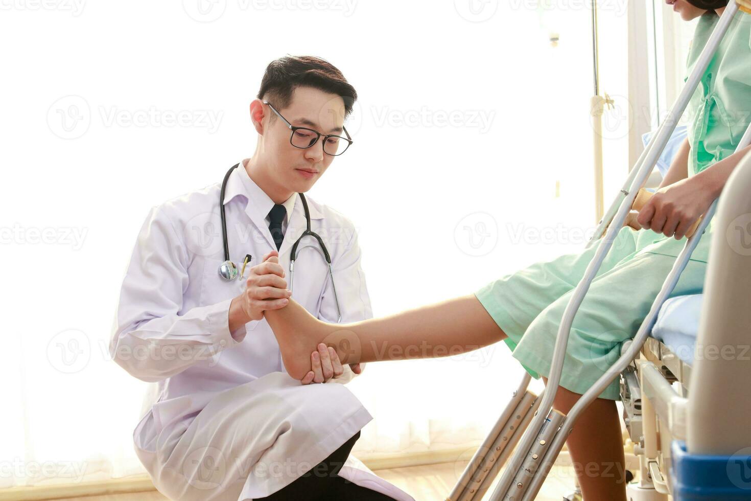 asiatico maschio medico l'esame il caviglia di un' femmina paziente seduta su un' assistenza infermieristica letto lei detiene un' canna per Aiuto sua camminare. il concetto di caviglia dolore, infiammazione, lesione. fisico terapia foto