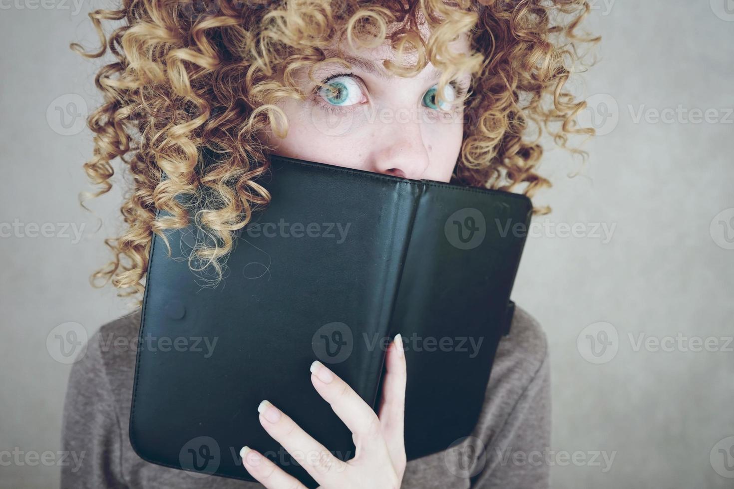 primo piano ritratto di una bella e giovane donna divertente con gli occhi azzurri e capelli biondi ricci dietro un'agenda o un ebook e lei è sorpresa foto