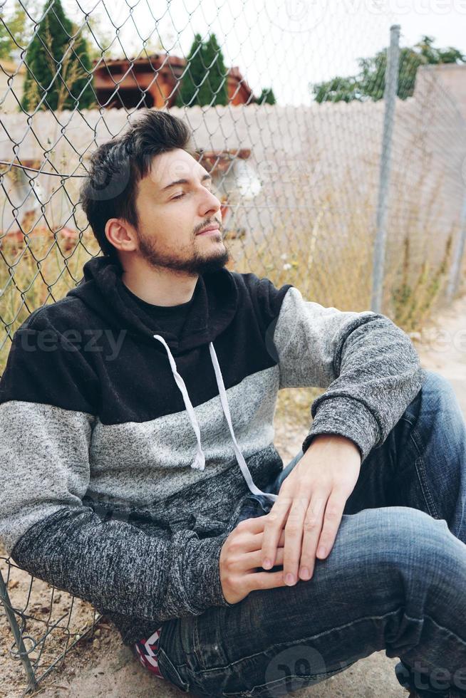 un giovane uomo attraente in calma seduto a terra e una recinzione all'aperto con gli occhi chiusi e la testa in alto foto