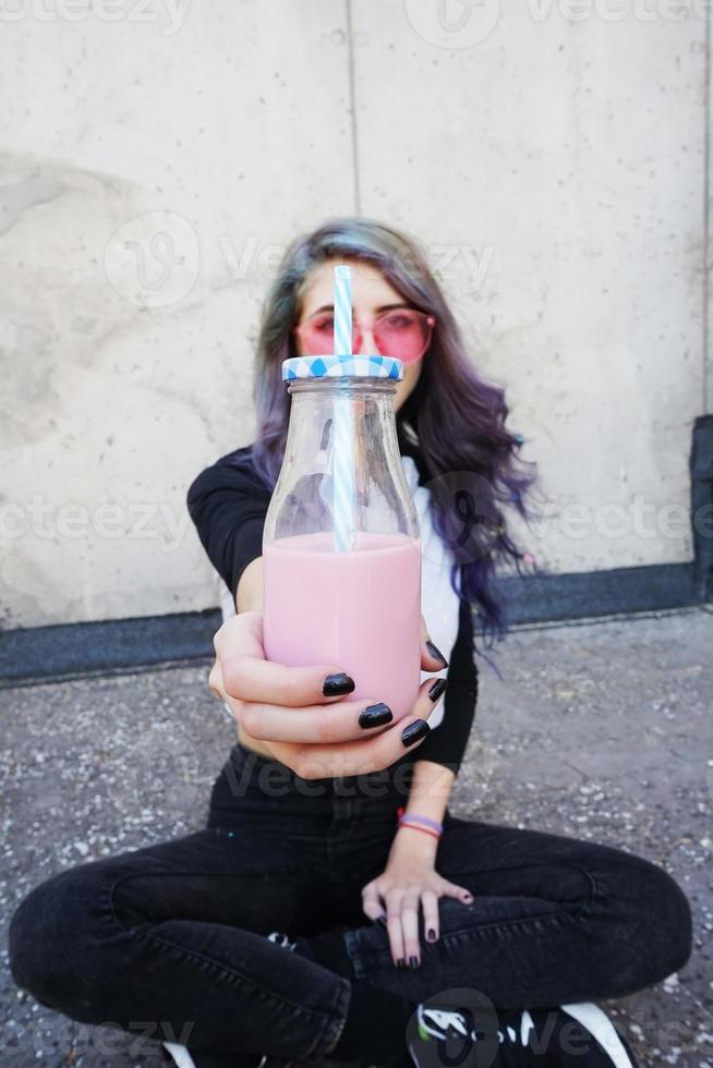 felice bella adolescente con occhiali da sole rosa esulta e si gode una bevanda rosa seduta su un terreno urbano foto