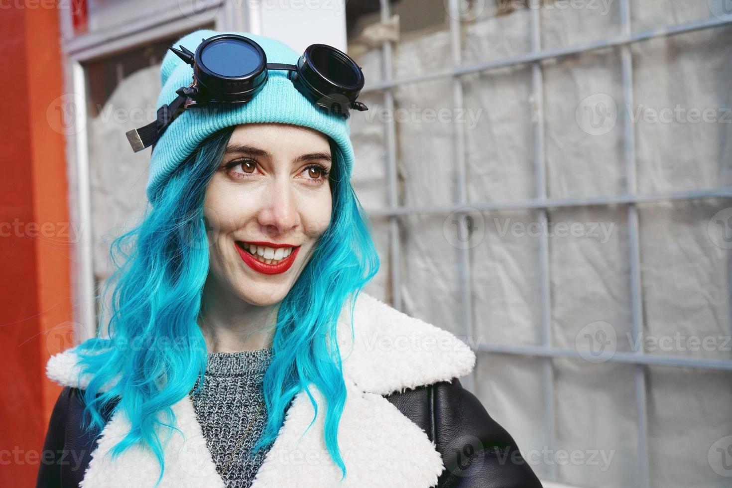 Ritratto di una giovane donna punk o gotica sorride con i capelli di colore blu e indossa occhiali steampunk neri e berretto di lana blu in una strada urbana all'aperto foto