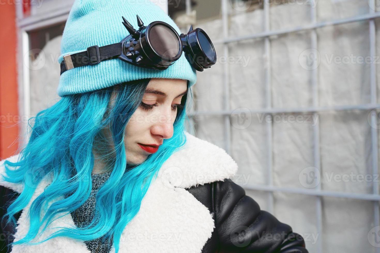 Ritratto di una giovane donna punk o gotica con capelli colorati di blu e indossando occhiali steampunk neri e berretto di lana blu in una strada urbana all'aperto foto