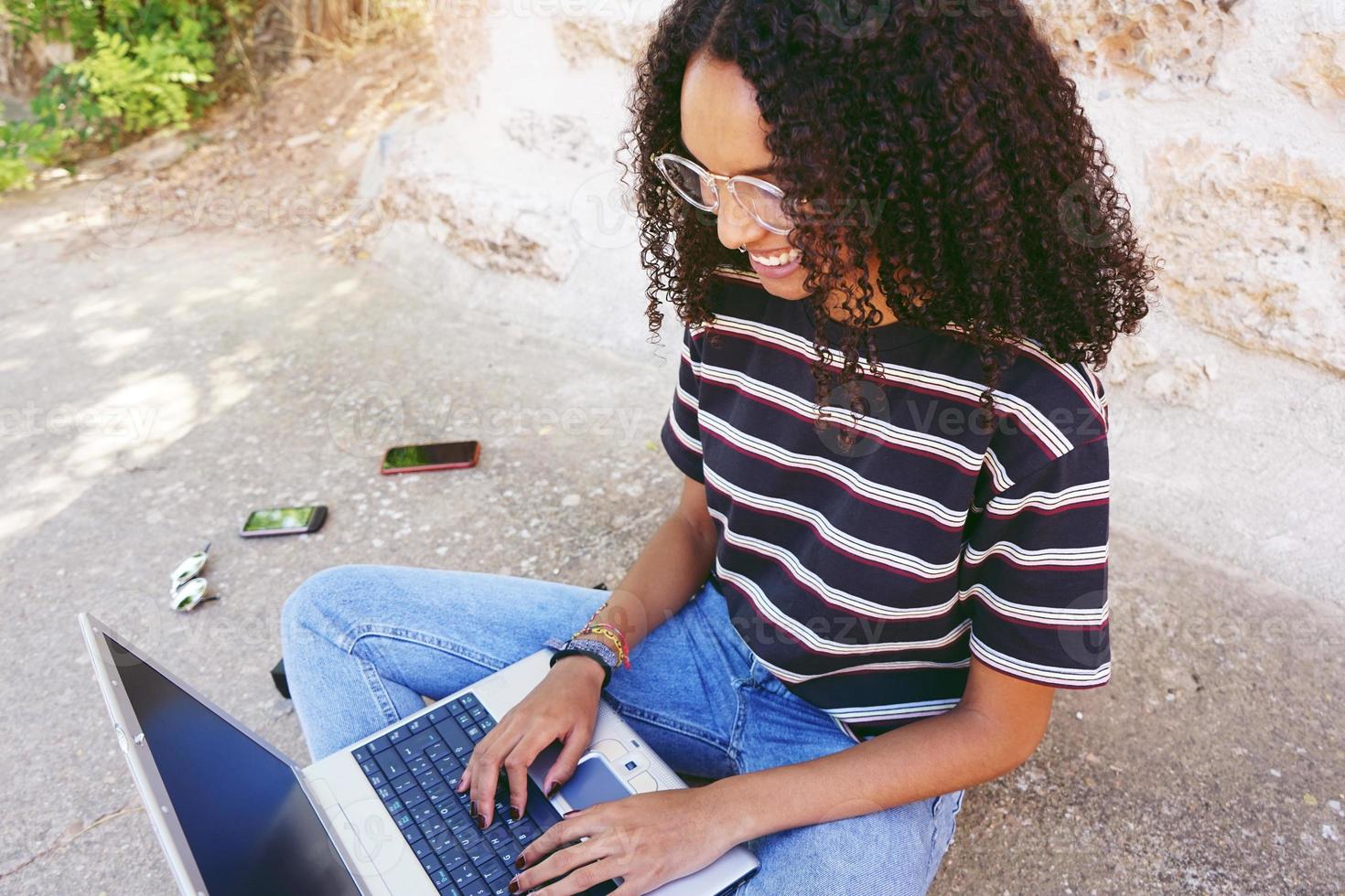 un ritratto di una giovane donna di colore sorridente felice con i capelli ricci che indossa occhiali, jeans e una t-shirt a righe, seduta per terra e lavora o fa i compiti foto