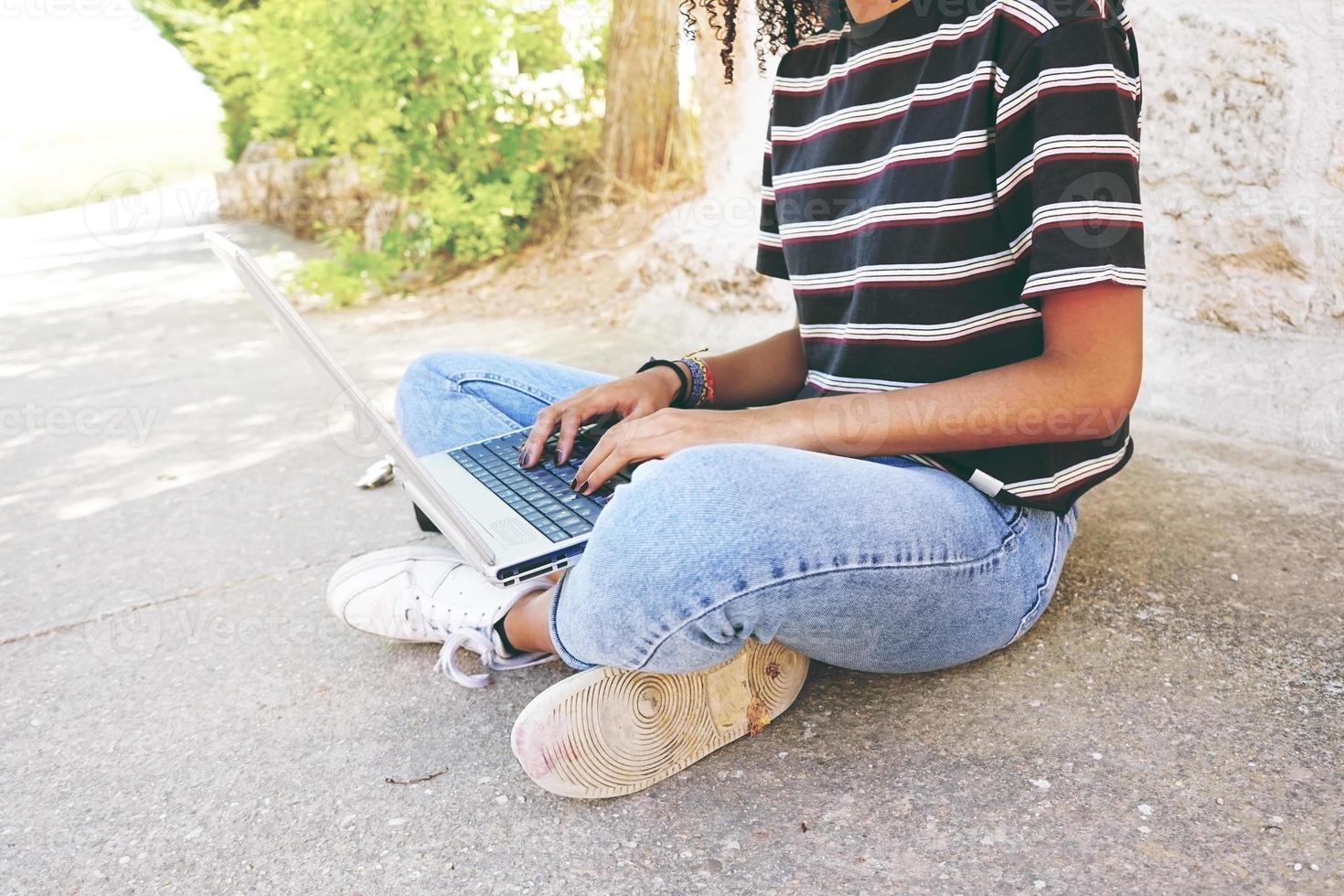 una giovane donna di colore con i capelli ricci che indossa jeans e una maglietta a righe, seduta per terra e lavora o fa i compiti foto