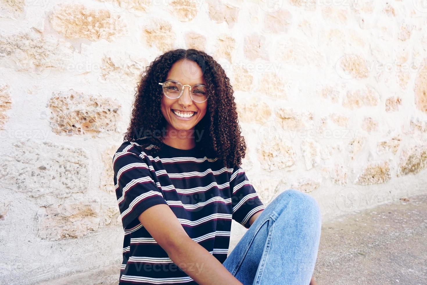 un ritratto di una giovane donna di colore sorridente felice che indossa occhiali, jeans e una t-shirt a righe, seduta per terra e capelli ricci foto