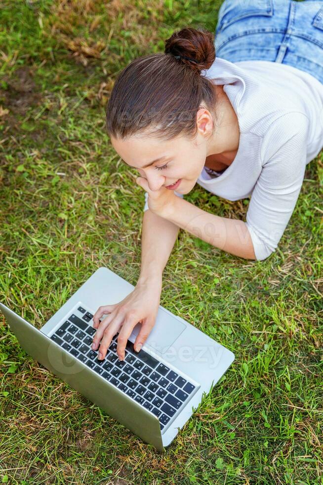 giovane donna dire bugie su verde erba prato nel città parco Lavorando su il computer portatile pc computer. libero professionista attività commerciale concetto foto