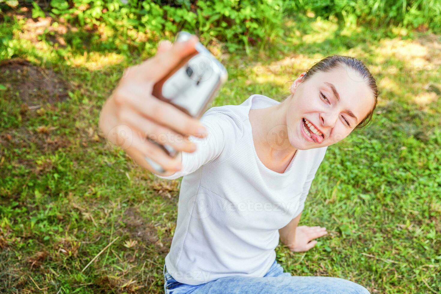 giovane ragazza prendere autoscatto a partire dal mani con Telefono mostrando lingua e divertente viso seduta su parco o giardino sfondo foto