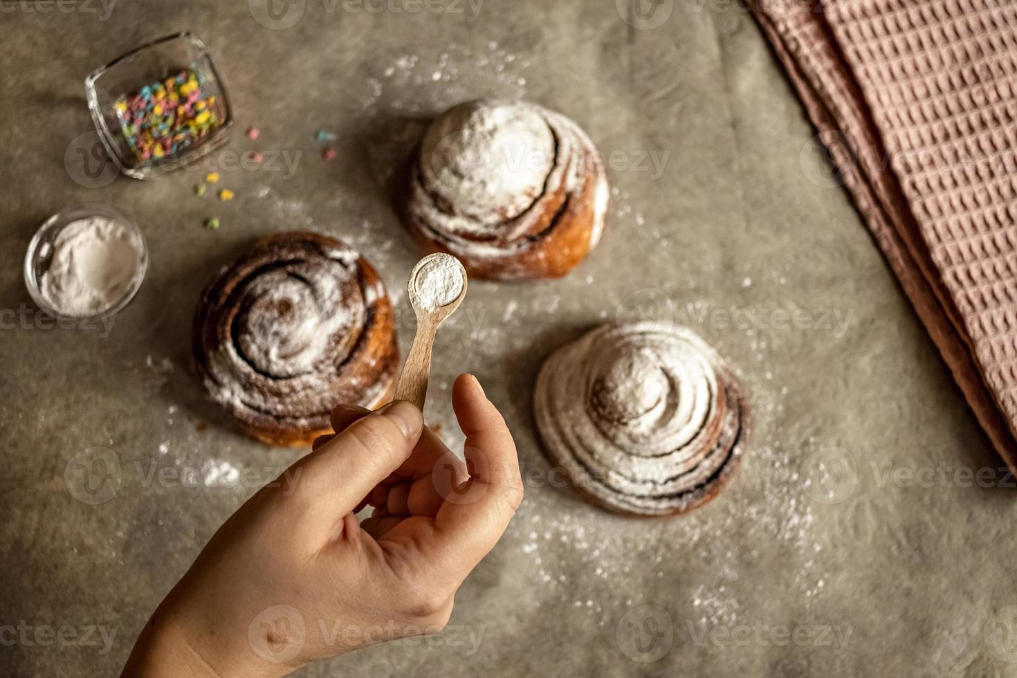 la mano della donna cosparge lo zucchero a velo sui panini alla cannella appena sfornati foto