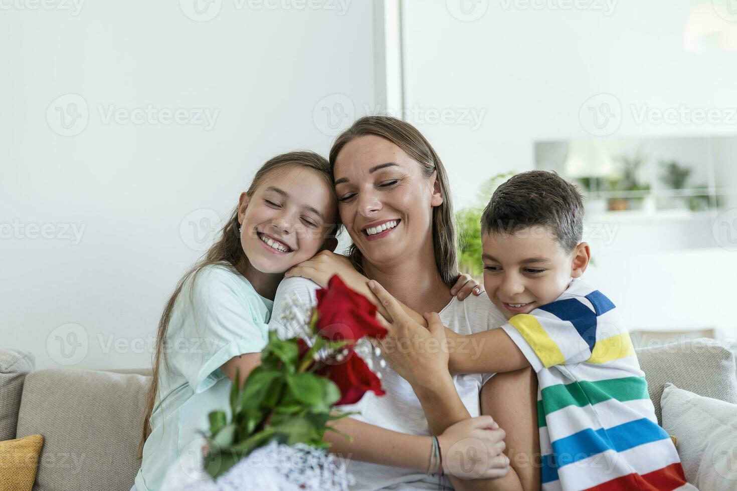 giovane madre con un' mazzo di Rose ride, abbracciare sua figlio, e allegro ragazza con un' carta si congratula mamma durante vacanza celebrazione nel cucina a casa foto