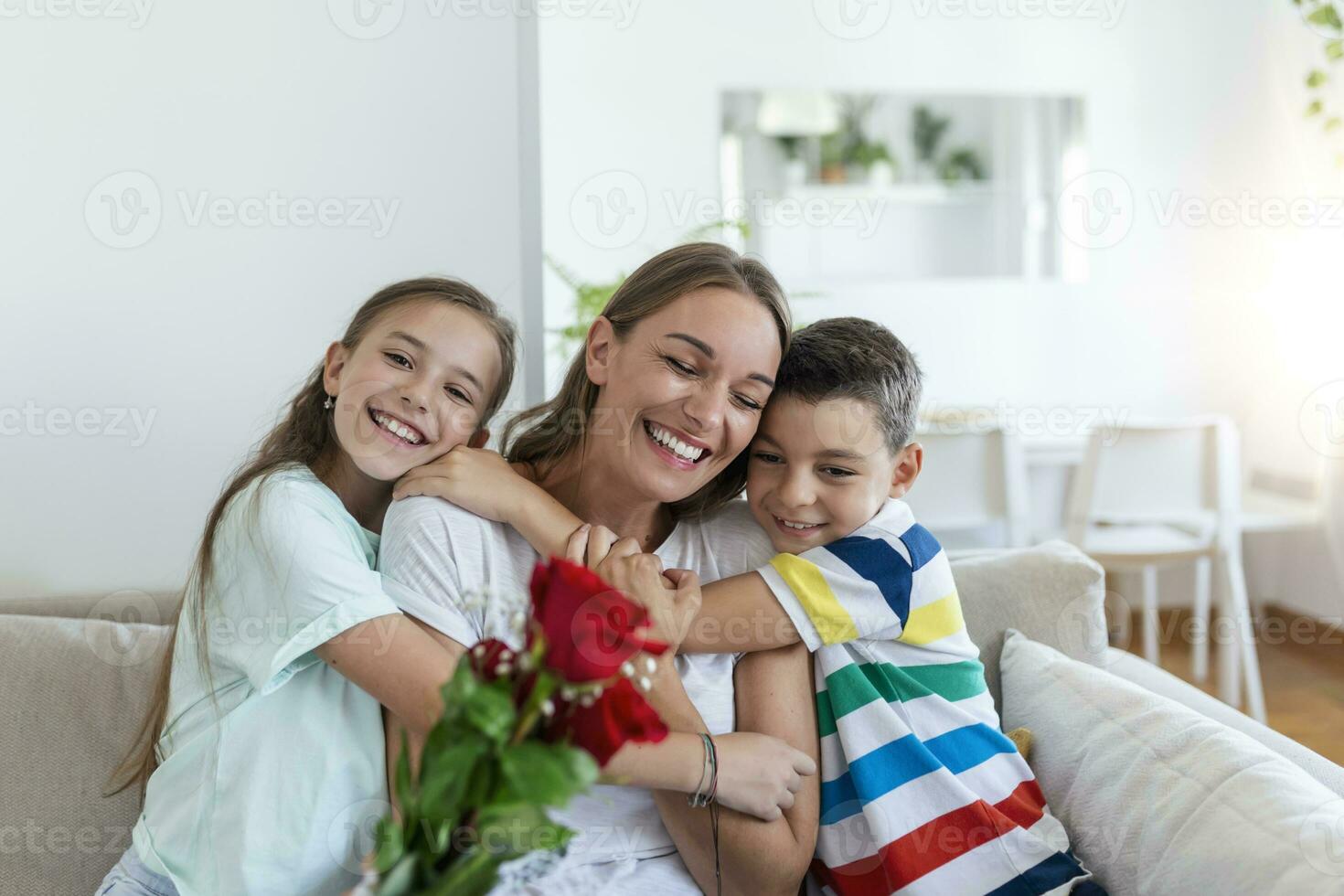 giovane madre con un' mazzo di Rose ride, abbracciare sua figlio, e allegro ragazza con un' carta si congratula mamma durante vacanza celebrazione nel cucina a casa foto