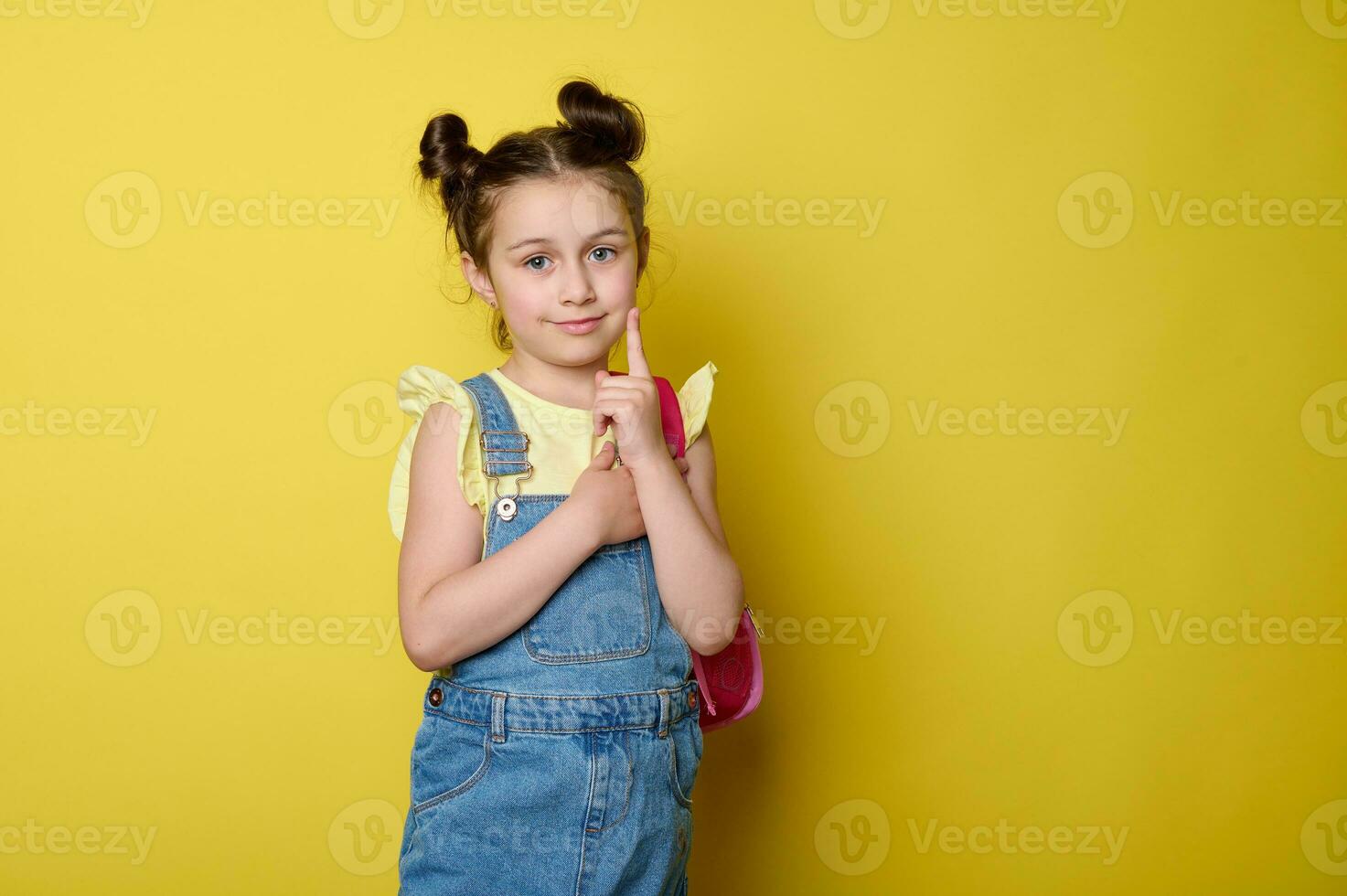 elegante sorridente contento primario scuola alunno ragazza nel casuale denim con rosa zaino, mostrando indice dito a telecamera foto
