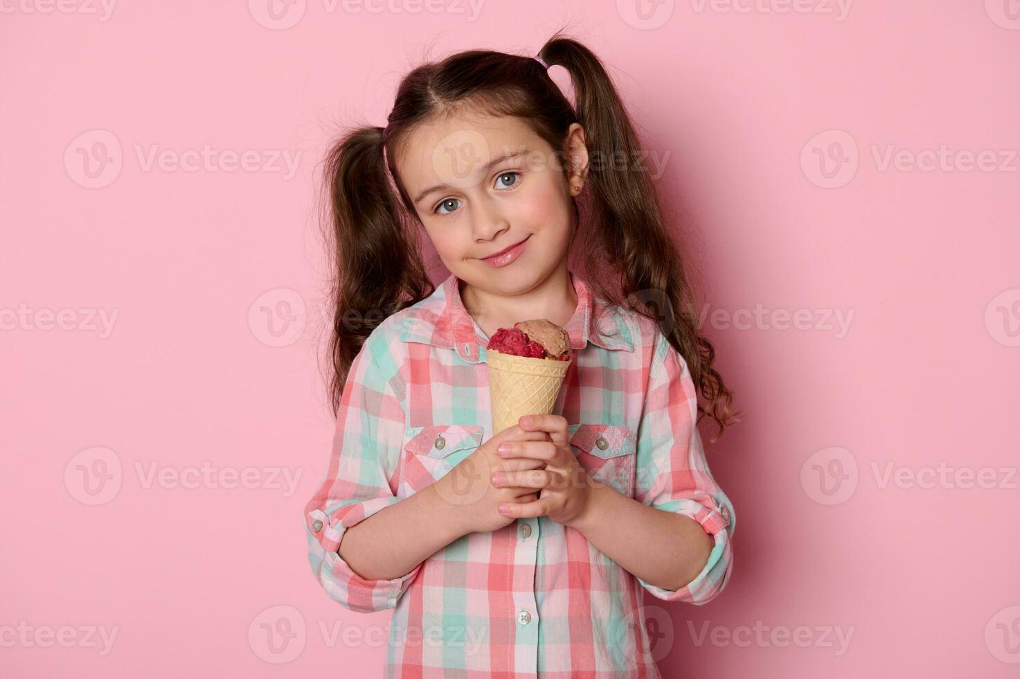 carino bambino ragazza con Due code di cavallo, sorrisi guardare a telecamera, Tenere delizioso ghiaccio crema, isolato su rosa sfondo. foto