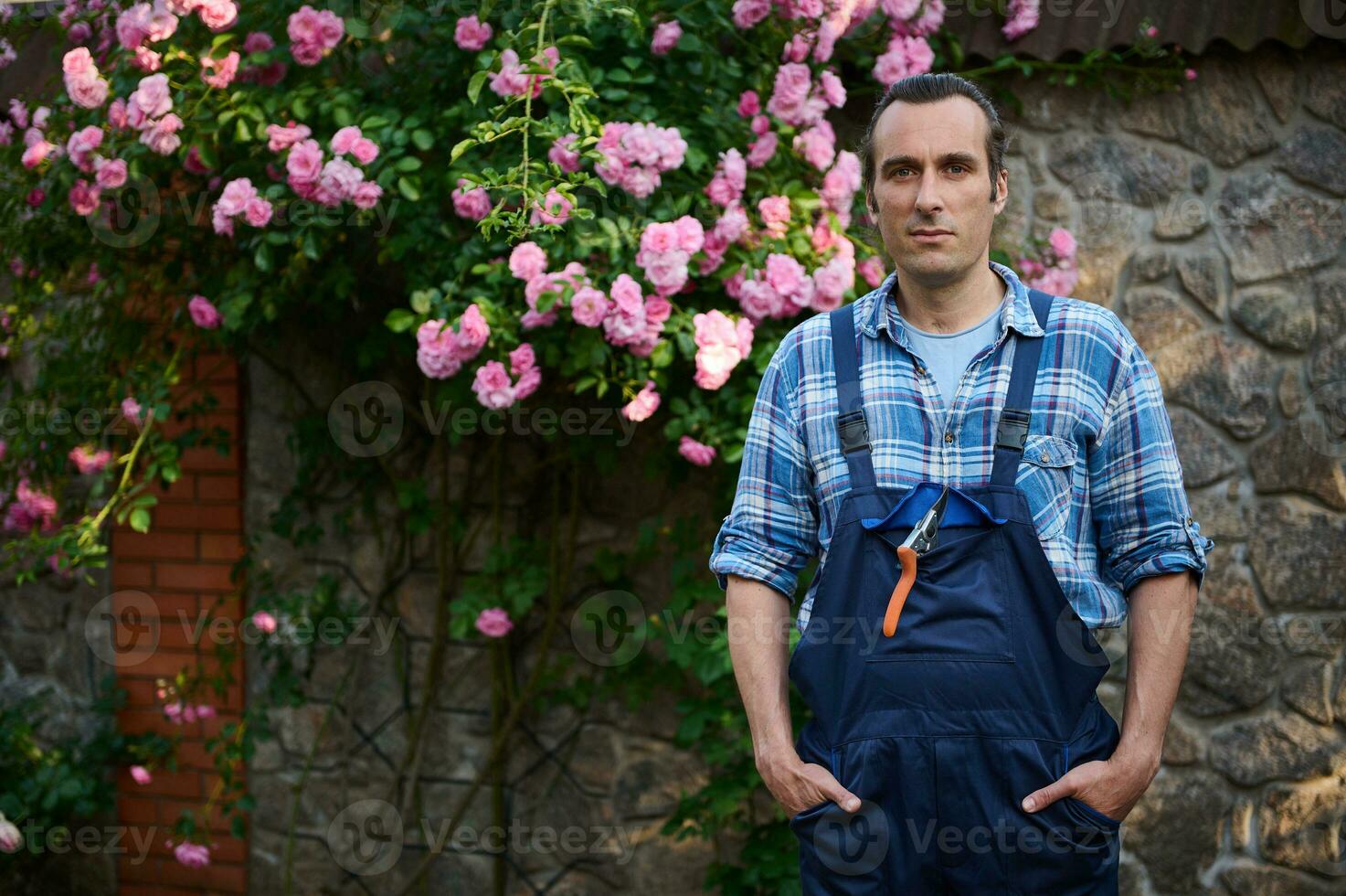 bello maschio giardiniere nel giardinaggio vestito, guardare a telecamera, in piedi a il suo posto di lavoro nel il Giardino dietro la casa di un' dimora foto