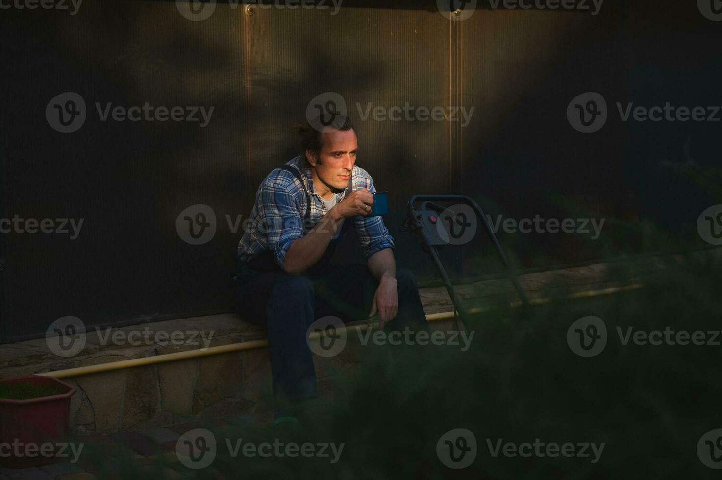 Visualizza attraverso siepe su il primo piano per oberati di lavoro uomo giardiniere con tazza di caffè, assunzione rompere dopo difficile Lavorando giorno foto