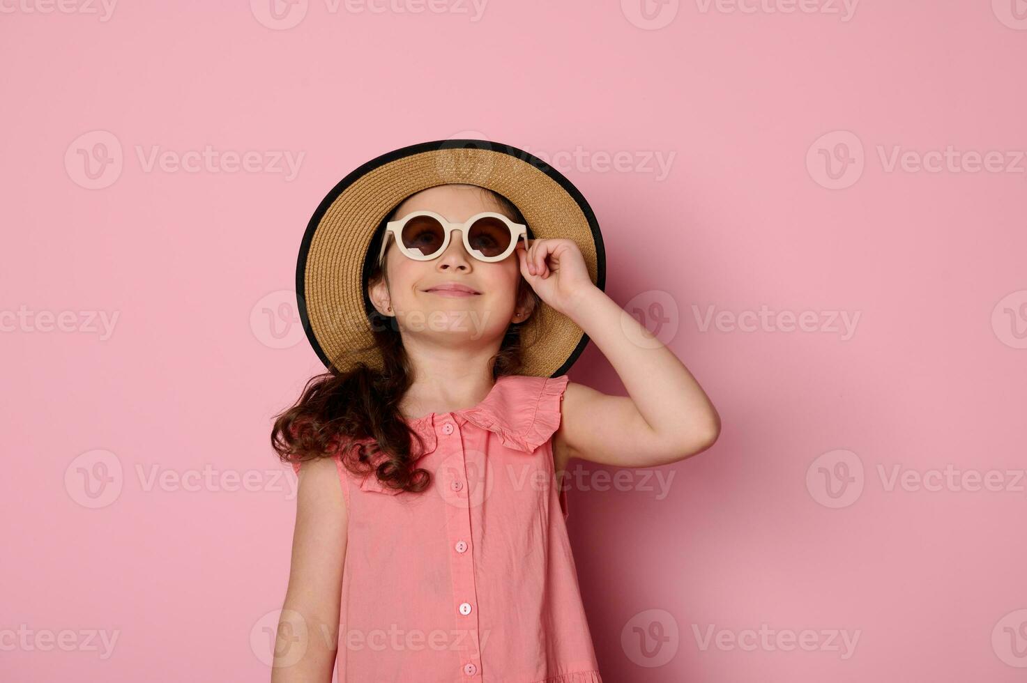 malizioso poco ragazzo ragazza indossare occhiali da sole, cannuccia cappello e estate prendisole, sorrisi guardare a telecamera isolato su rosa foto