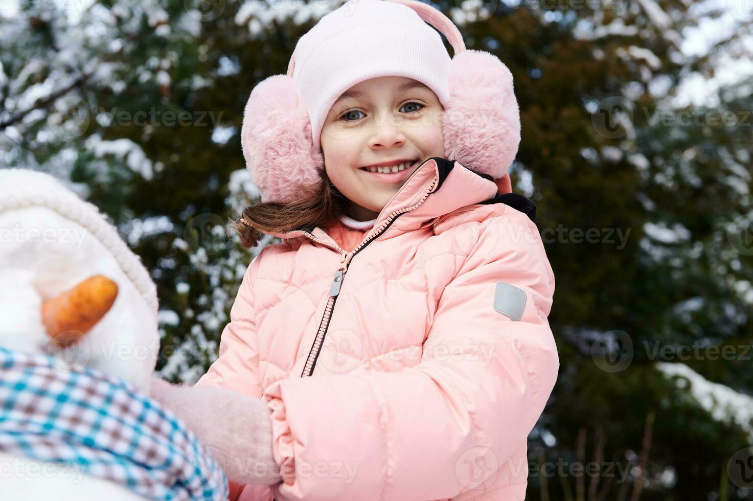 autentico bellissimo contento poco bambino ragazza giocando con un' pupazzo di neve su un' nevoso inverno parco, sorridente guardare a telecamera foto