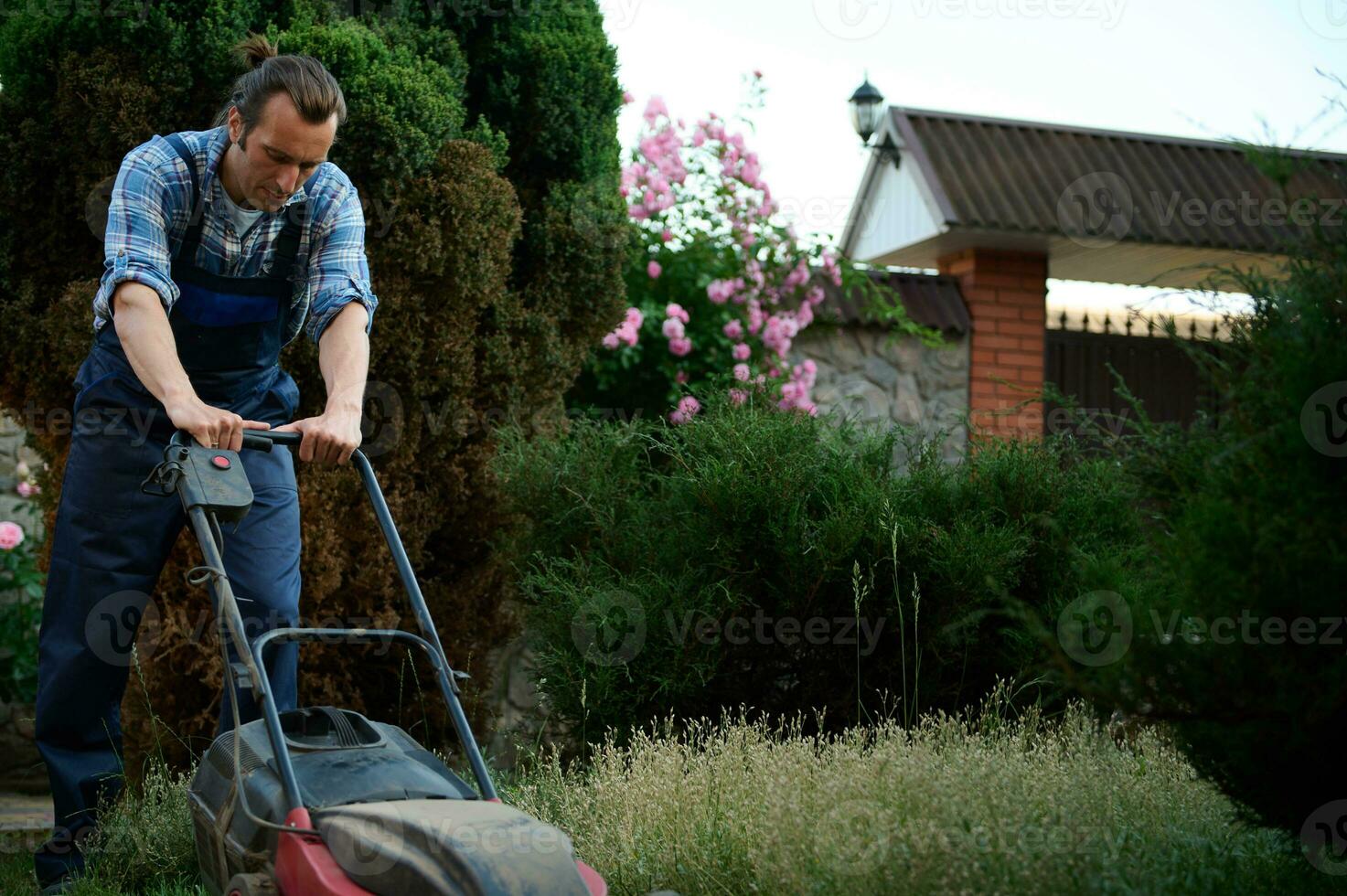 ispanico giardiniere nel scacchi blu camicia e opera giardinaggio uniforme, falcia prato utilizzando elettrico prato falciatore nel Giardino dietro la casa foto