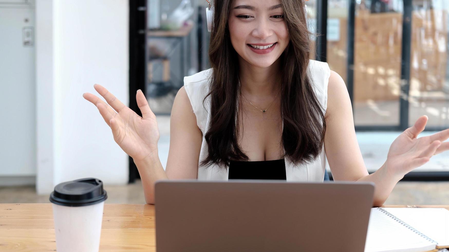 giovane donna d'affari asiatica che indossa le cuffie che studia online guardando un podcast webinar sul laptop, ascoltando e imparando la chiamata in conferenza del corso di istruzione, prendere appunti seduti a una scrivania, concetto di e-learning foto