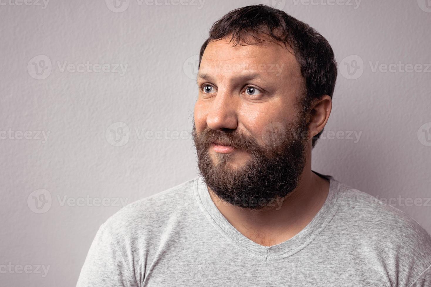 uomo barbuto felice in maglietta grigia che guarda lontano mentre sta in piedi contro il muro grigio foto