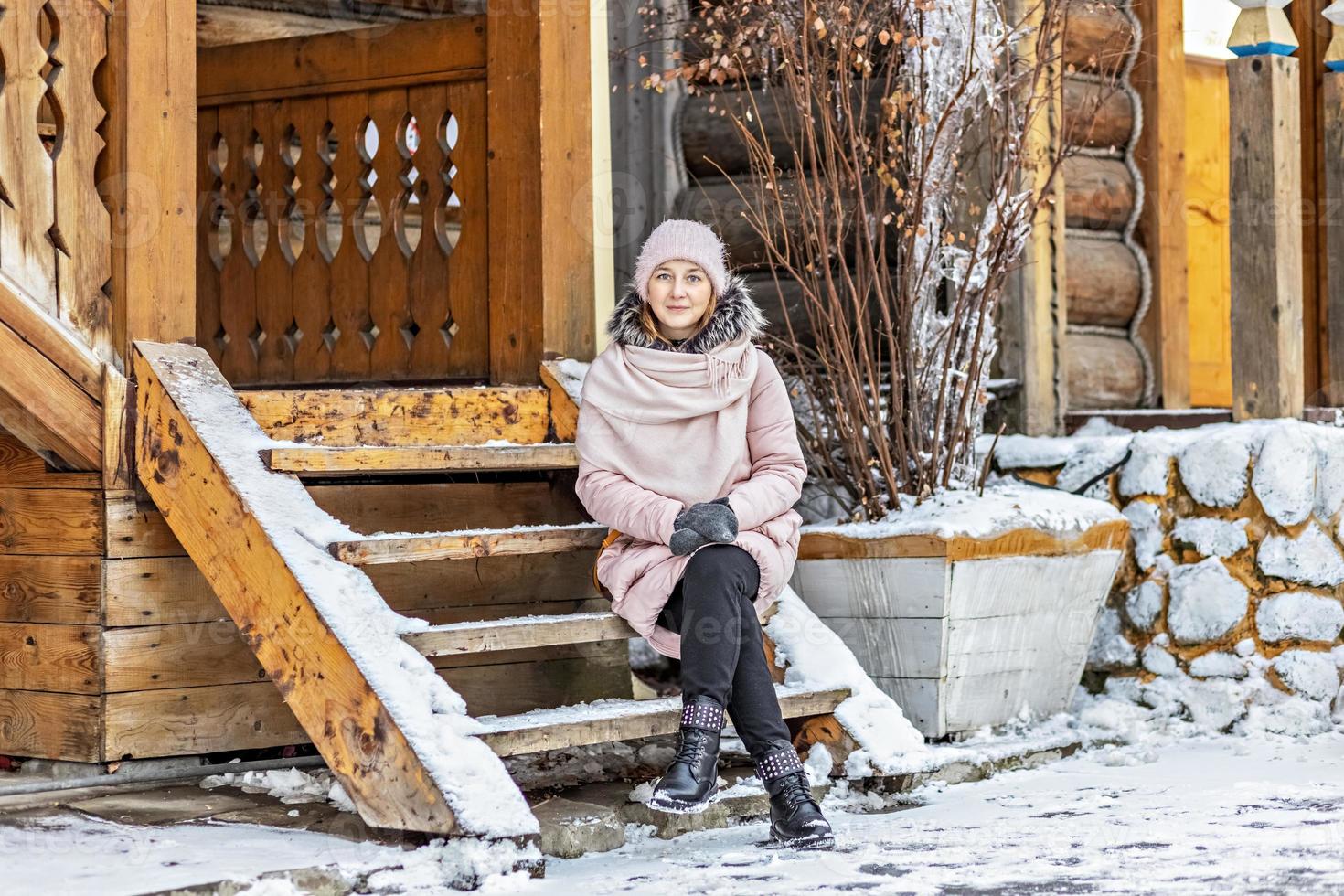 giovane donna vestita calorosamente in posa sotto il portico di una casa in legno nel villaggio. vacanze invernali in campagna foto