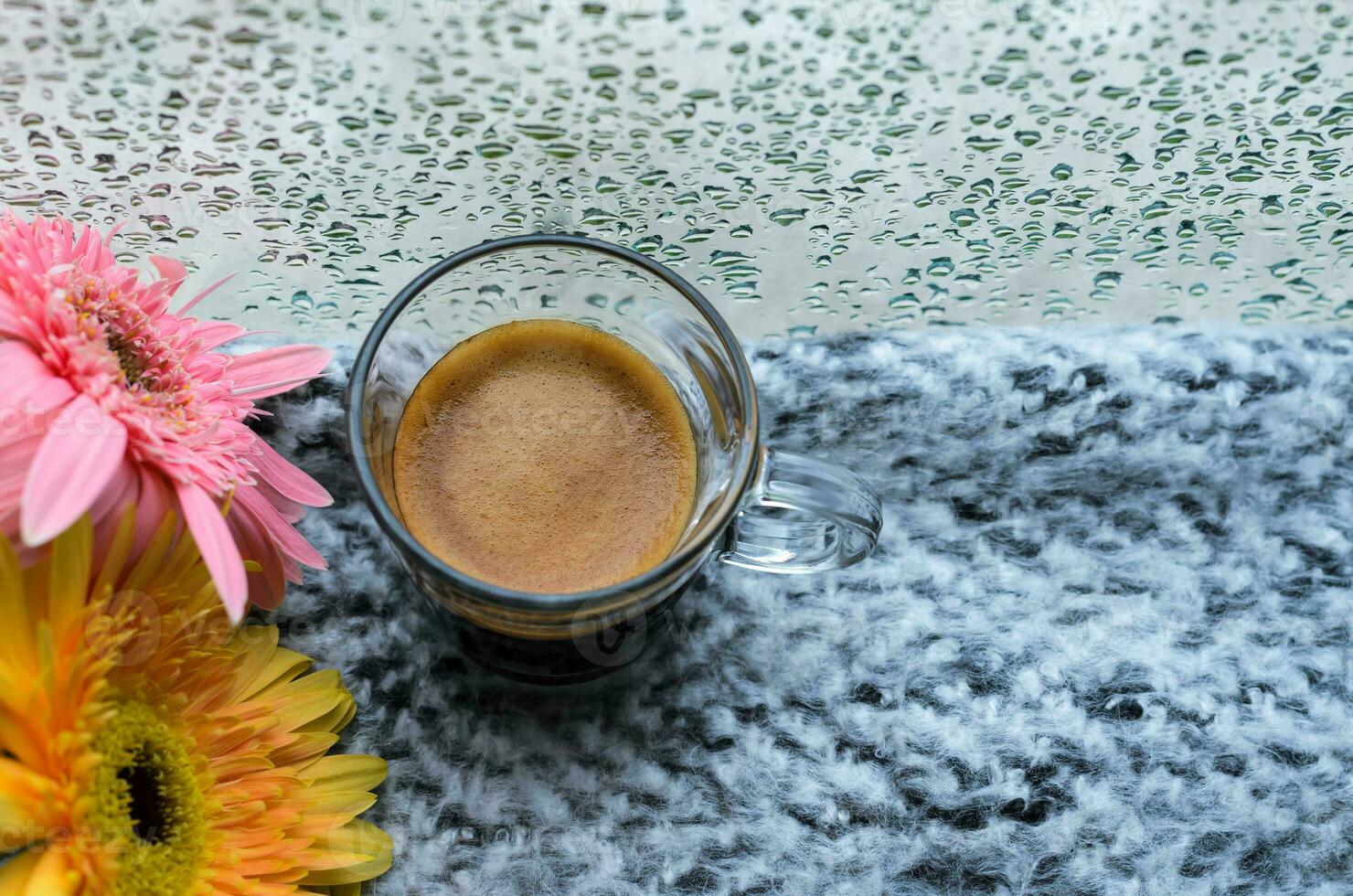 un' bicchiere di caffè espresso caffè tiro mette su letto con gerbera fiori quando pioggia far cadere su finestra nel pioggia giorno. foto