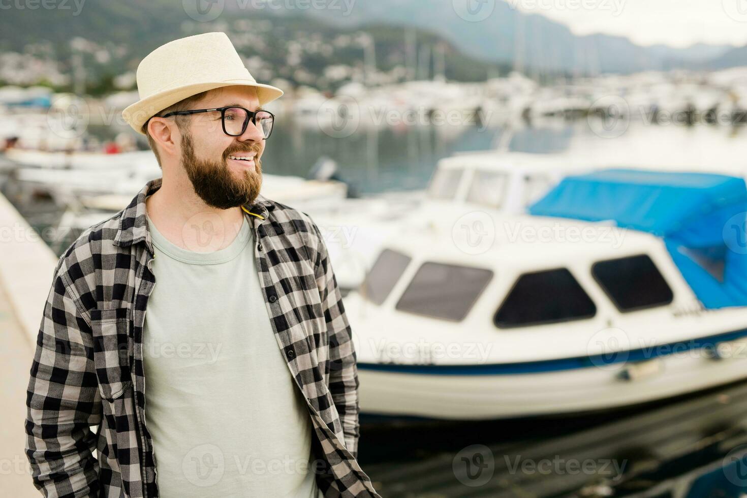 bello uomo indossare cappello e bicchieri vicino marina con yacht. ritratto di ridendo uomo con mare porta sfondo con copia spazio foto