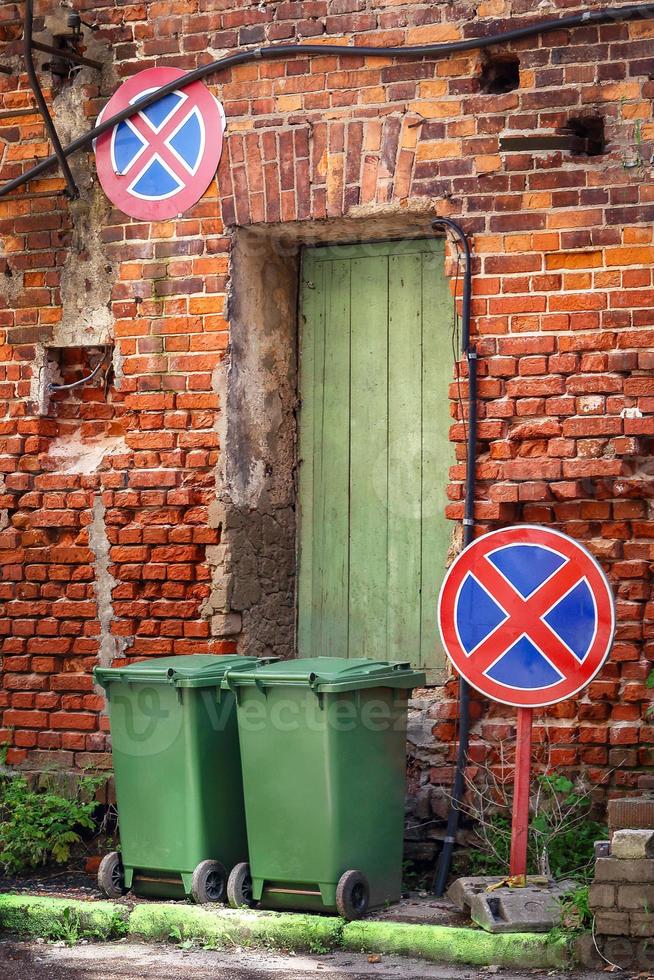 due bidoni della spazzatura in piedi sulla strada con un muro di mattoni rossi con una porta di legno verde e due segnali stradali restrittivi foto