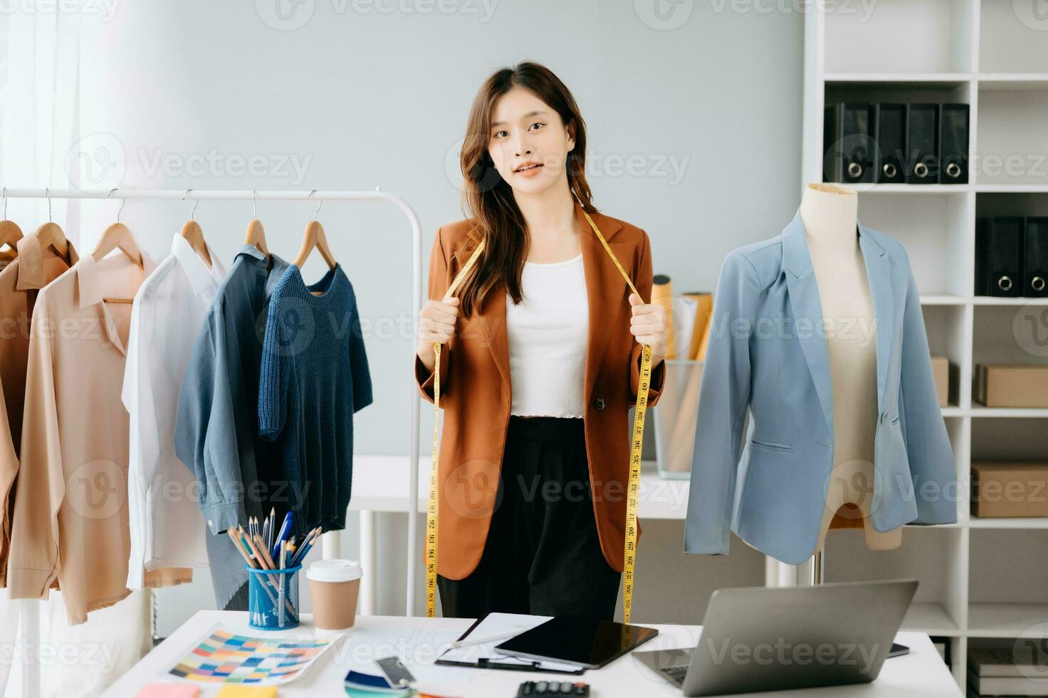 moda progettista donna parlando inteligente Telefono e utilizzando il computer portatile con digitale tavoletta computer nel moderno studio il Abiti sospeso su il cremagliere mattina leggero foto