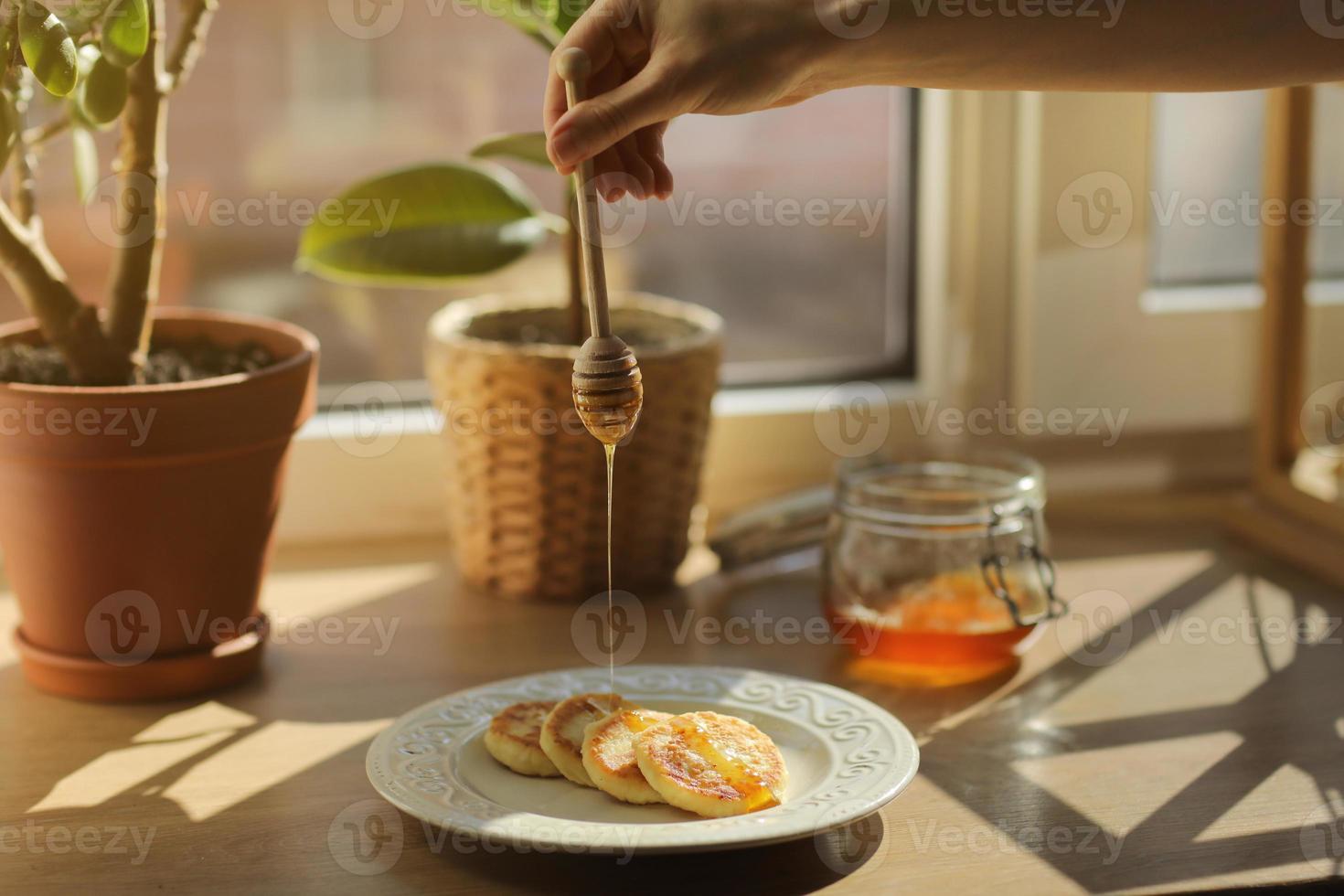 cheesecake o syrniki su un piatto con miele, sullo sfondo un vasetto con miele e ficus. una donna che versa un cucchiaio di legno per il miele che scorre miele. avvicinamento. messa a fuoco selettiva foto