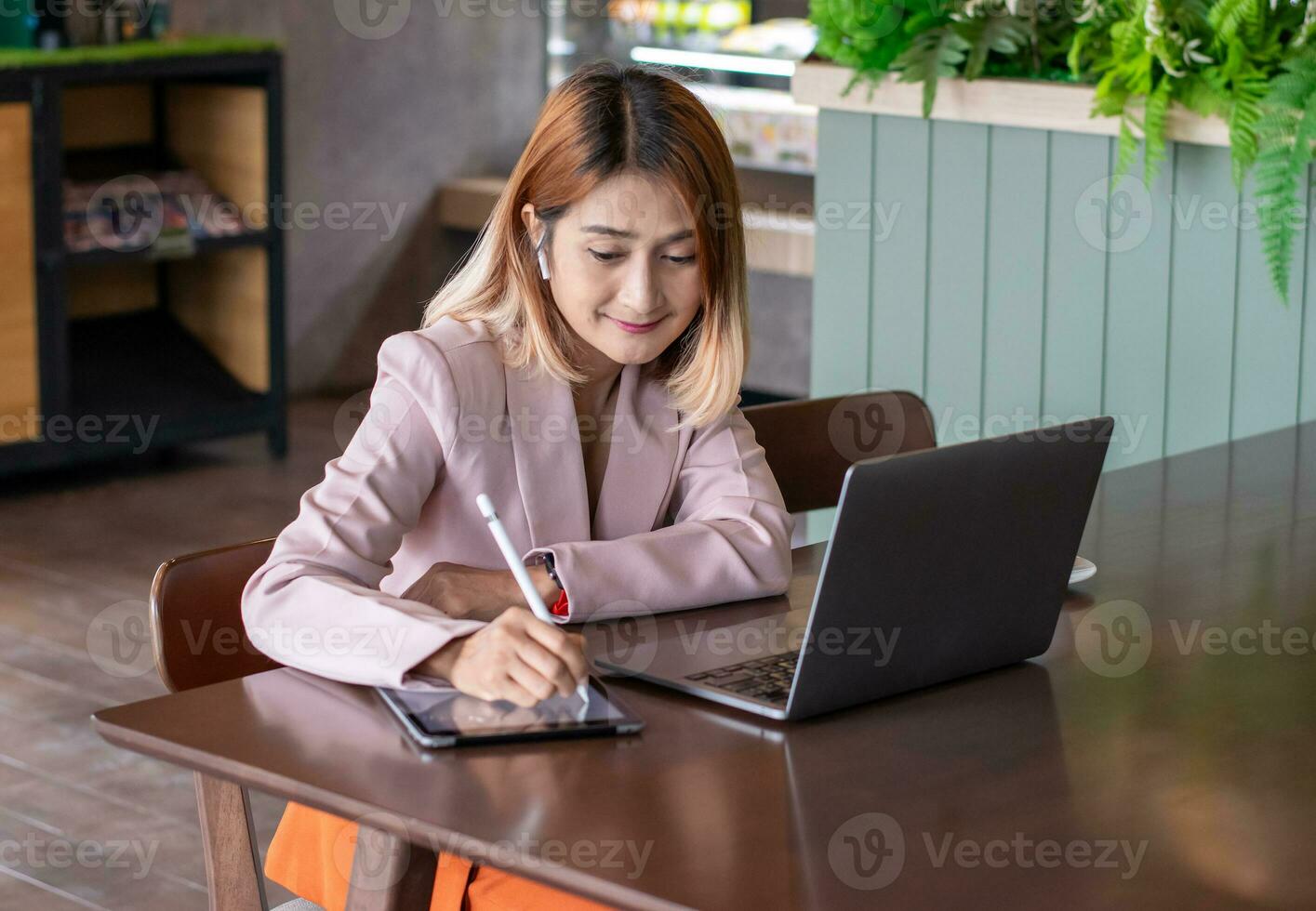 donna d'affari indossare un' completo da uomo rosa seduta a il caffè negozio e utilizzando il computer portatile conferenza video chiamata per parlare in linea incontro e utilizzando tavoletta matita per Scrivi rapporto su contabilità finanza attività commerciale. foto