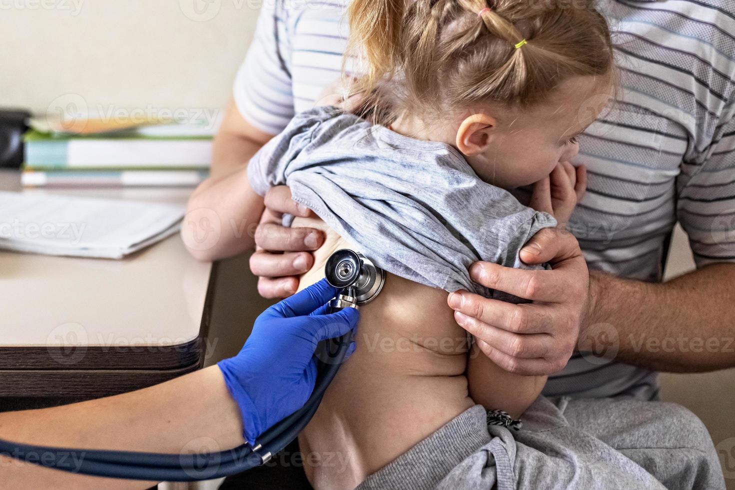 bambina tra le braccia di suo padre nell'ufficio del medico presso la clinica. il medico esamina il bambino, ascolta i polmoni con un fonendoscopio. trattamento e prevenzione delle infezioni respiratorie. foto