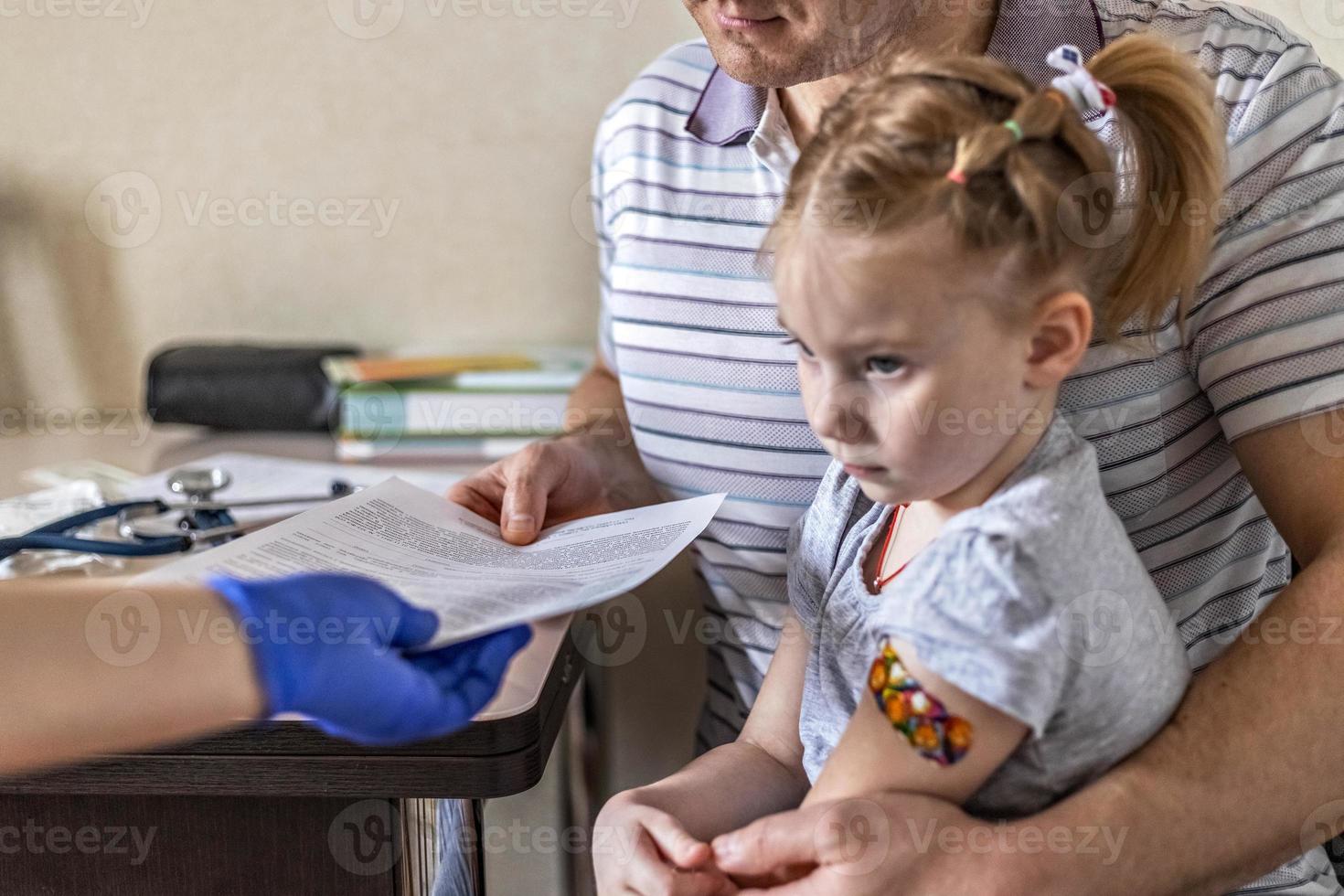 bambina con suo padre nell'ufficio del medico presso la clinica. vaccinato contro il coronavirus. il dottore passa i documenti al papà della ragazza. il concetto di vaccinazione da covid-19 foto