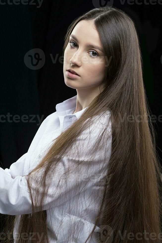 bellissimo giovane ragazza con lungo dritto capelli nel un' bianca camicia in posa nel il studio su un' grigio sfondo foto