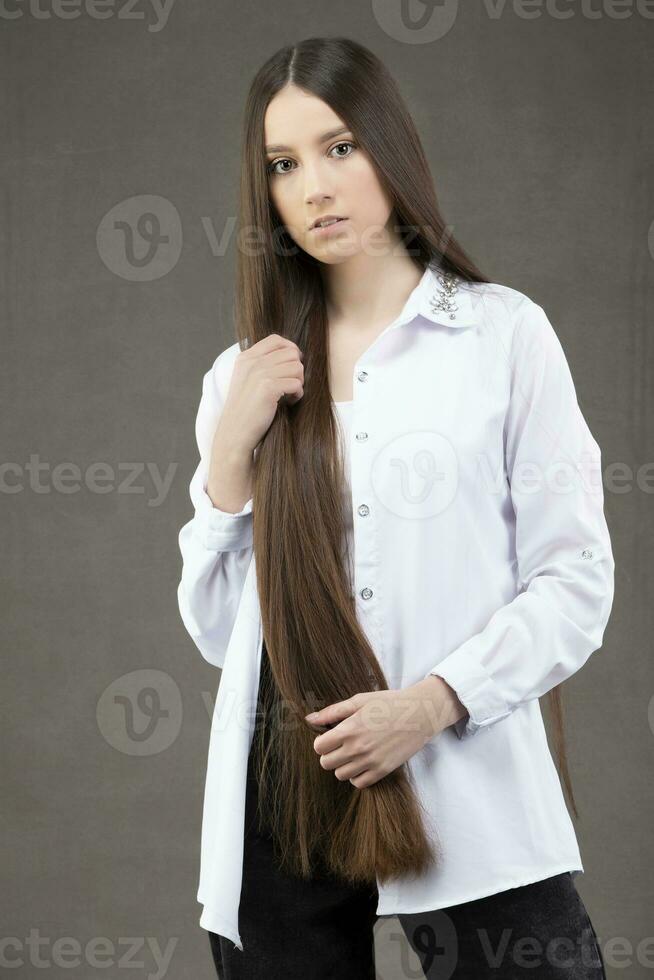 bellissimo giovane ragazza con lungo dritto capelli nel un' bianca camicia in posa nel il studio su un' grigio sfondo foto