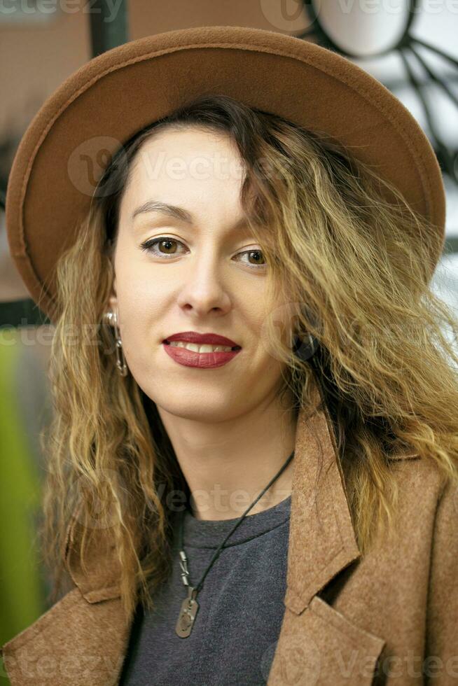 un' bellissimo ragazza nel un' beige elegante cappello con lungo Riccio capelli sembra a il telecamera e sorrisi. foto