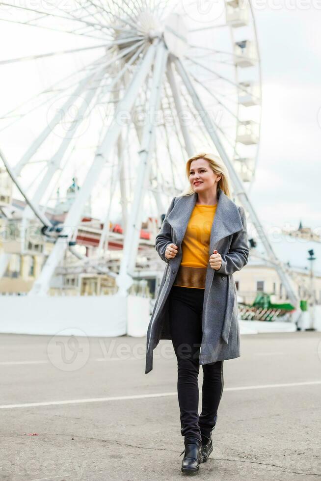 elegante signora a piedi in giro città. all'aperto foto di bella ragazza con affascinante Sorridi in posa nel grigio cappotto su urbano sfondo Ferris ruota