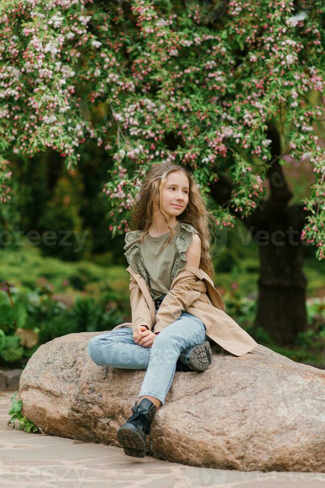 carino adolescenziale ragazza con lungo Marrone capelli nel impermeabile è seduta su un' roccia nel il parco foto